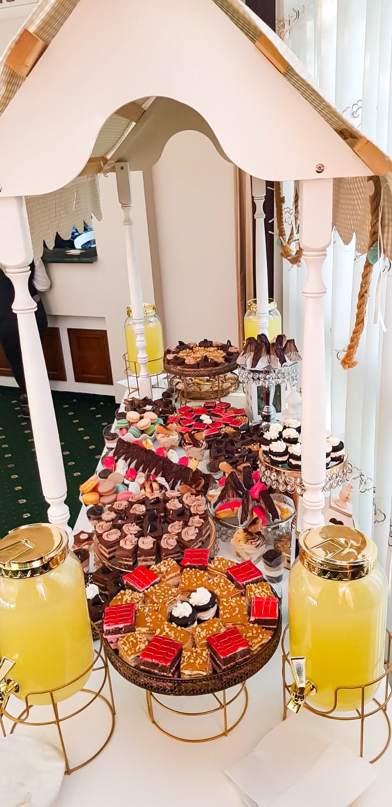 Masă dulce de eveniment pregătită de Cofetăria Giumbo, aranjată sub o arcadă albă, cu limonade și prăjituri asortate